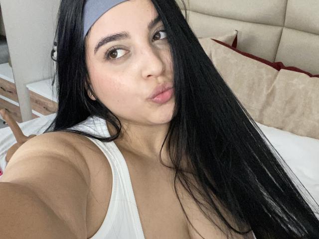 ShinyMurpy with long black hair, pouty lips, and a grey headband, wearing a white tank top, showing her tits, taking a selfie in a bedroom.