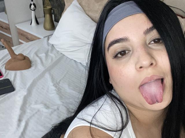 ShinyMurpy with long black hair, gray headband, brown eyes, and tongue out, showing a pink tongue piercing, lying on a bed with a dildo in the background.
