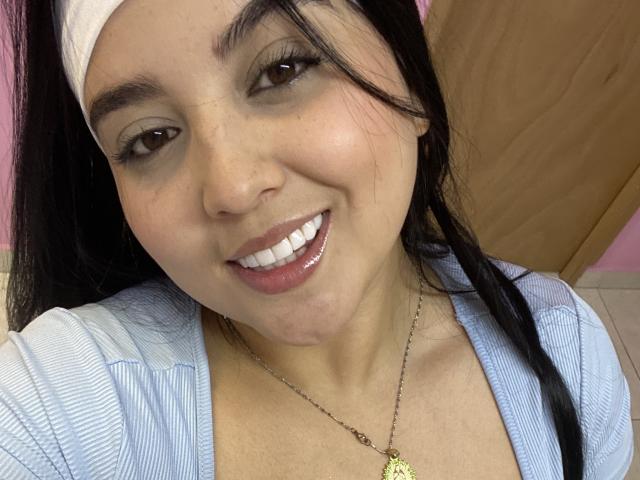 ShinyMurpy has medium brown skin, black hair, and a white headband. She's smiling with glossy pink lips, wearing a blue top, and a gold necklace with a small pendant.
