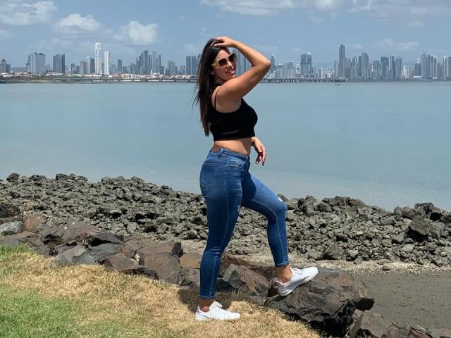 RachelWildest with dark hair, black tank top, blue jeans, white sneakers, standing on rocks, one hand on head, waterfront city skyline, clear blue sky, green grass, sunny day.