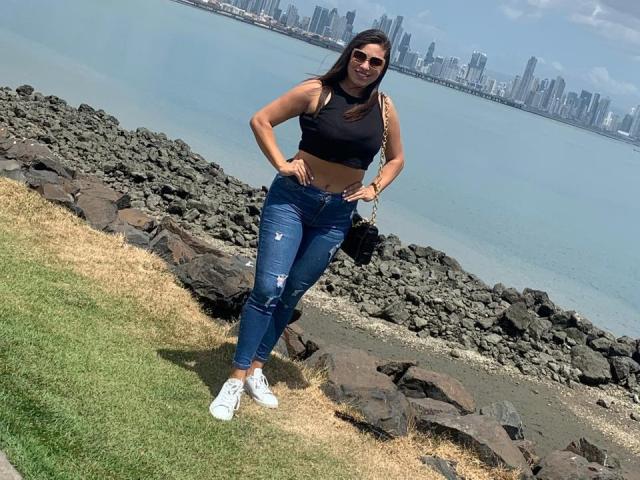 RachelWildest, with medium tits, wearing a black crop top, blue ripped jeans, white sneakers, standing on grass, rocks, and water, smiling, hands on hips, city skyline in background.