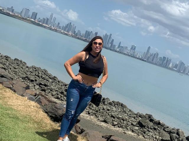 RachelWildest with dark hair, sunglasses, black crop top, and ripped blue jeans, standing on rocky shore, hands on hips, city skyline and blue water in background.