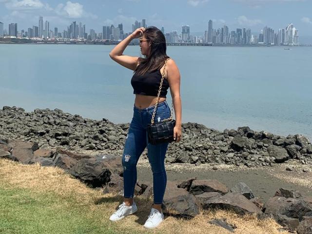 RachelWildest with medium tits, wearing a black crop top, ripped blue jeans, white sneakers, and a black purse, standing by rocky water, hand on head, city skyline in background.