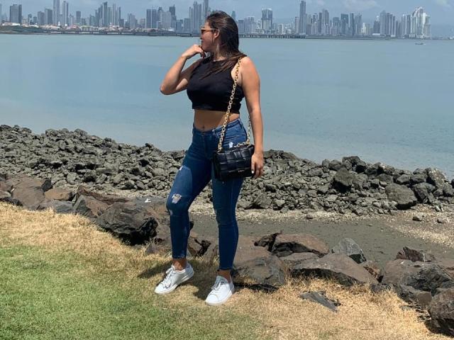 RachelWildest with tanned skin, black crop top, ripped blue jeans, white sneakers, black purse, standing on grass, rocks, and water, city skyline in background, eating or talking.