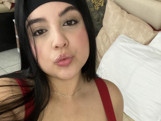 ShinyMurpy, avec de longs cheveux noirs, des yeux marron, des lèvres roses, portant un haut rouge et un bandeau noir, est allongée sur un lit avec des oreillers blancs. Elle porte un collier en argent..
