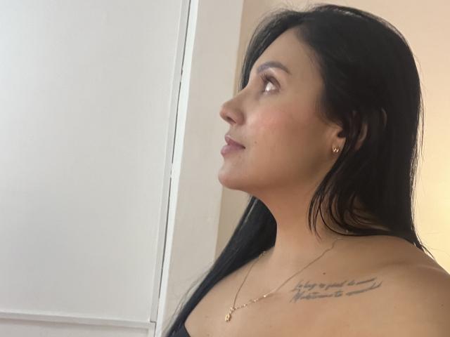 YessiJaymes has long black hair, looking to the side with a slight pout, wearing a gold necklace, and has a tattoo on her right shoulder. Her ass is not visible.