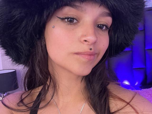 MarietteJones has a cute face with black fur hat, nose piercings, and winged eyeliner. She's got long brown hair, and a subtle smile. Her tits aren't visible.
