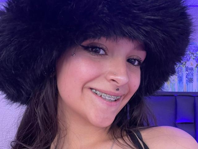 MarietteJones with dark fur hat, black eyeliner, nose piercings, and glittery lip piercings, smiling at the camera. She has light skin and brown hair.