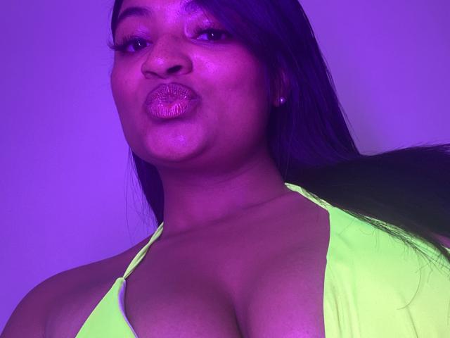 CameronJones with purple-lit face, pouty lips, and neon green halter top showing her big tits. Long black hair, subtle makeup, earrings, and a confident expression.