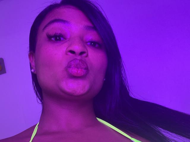 Cameron Jones, una mujer de piel oscura con cabello negro largo, labios gruesos, vestida con un top de tirantes verde neón, rostro iluminado en morado, pequeños aretes de perlas, mirando a la cámara, en un selfie..