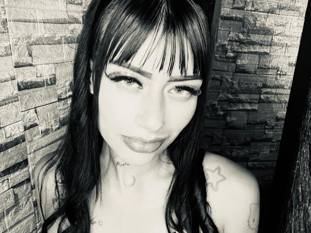 Black-and-white photo of JeffAndRechell with dark hair, bangs, long eyelashes, tattoos, and a subtle smile. She's looking up with a slight smirk, showing her tits.