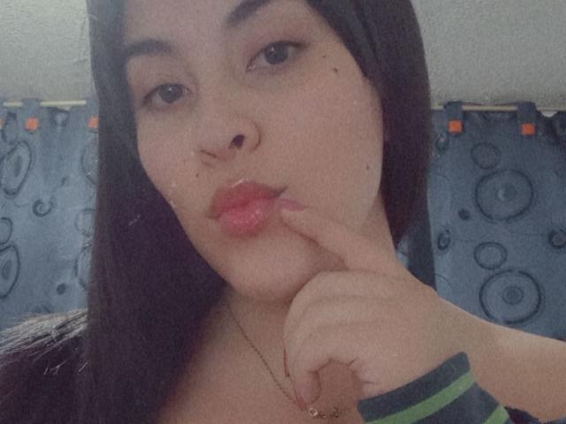 SelenaF has pouty pink lips, her finger slightly touching her lower lip, dark brown hair, and a gold necklace. She's wearing a green and gray striped wristband.
