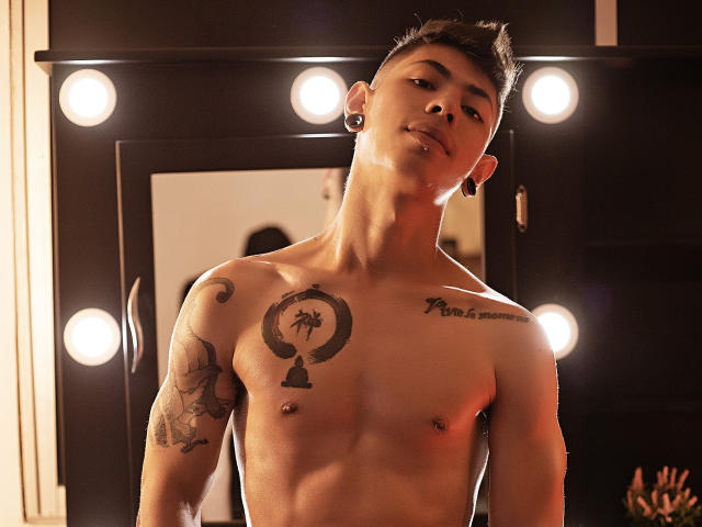 Shirtless ArturCarter with tattoos, dark hair, and ear gauges, standing in front of a mirror with round lights, looking down with a slight smile, showing a nipple.