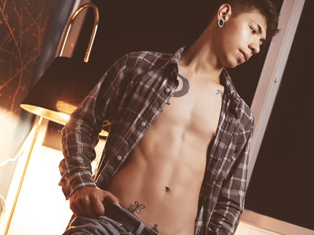 ArturCarter, shirtless, plaid, unbuttoned, showing abs, ear gauges, crescent tattoo, side profile, lowering jeans, tattoo on lower belly, short brown hair, dim light, slight smile.