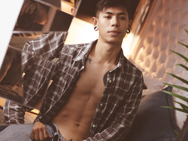 ArturCarter, shirtless with a plaid button-up, has a tattoo on his chest, black ear gauges, and a slight nipple piercing, sitting in a relaxed, slightly seductive pose.