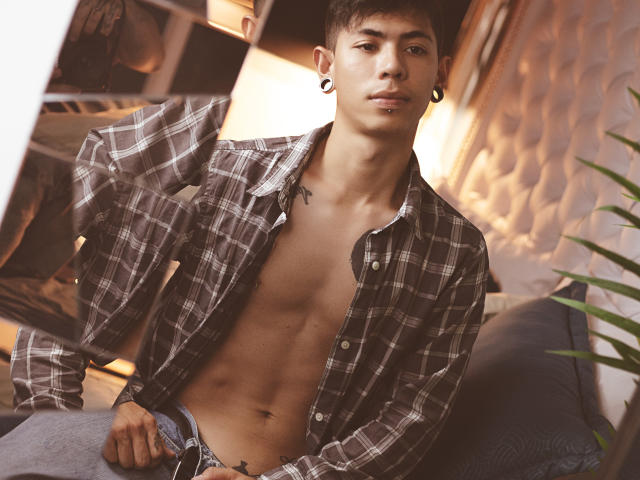 ArturCarter with a shirt open, revealing a toned, muscular, shirtless chest and abs, wearing unbuttoned jeans, ear gauges, and a nose ring, sitting indoors.