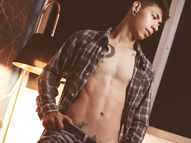 ArturCarter, shirtless, with a toned abs, plaid button-up, ear gauges, moon tattoo, and flower tattoo, holding pants, under warm lamp, looking right, short dark hair, night.