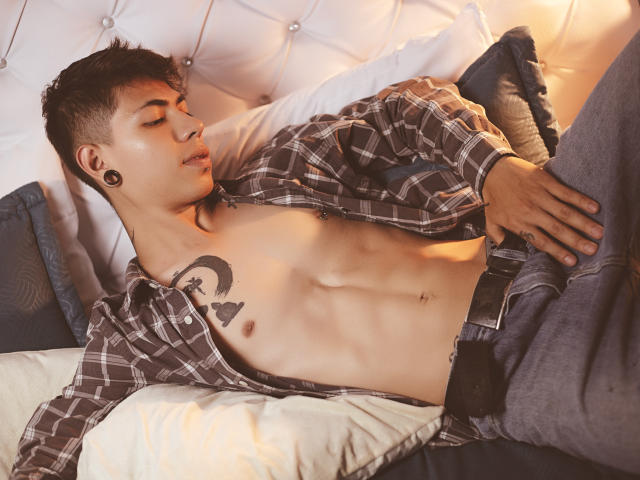 ArturCarter with a toned, shirtless body, plaid shirt open, blue jeans, black ear gauges, and a moon tattoo on his right chest, lying on a bed.