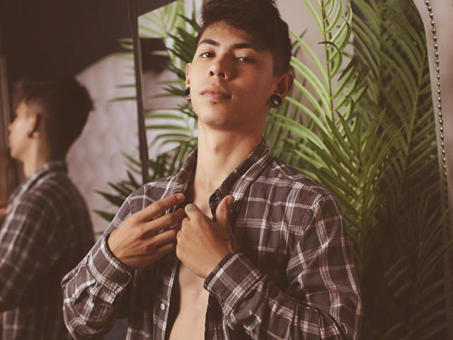 ArturCarter with short dark hair, small nose ring, and plugs, wearing an unbuttoned plaid shirt, revealing a bare chest, standing in front of a mirror.