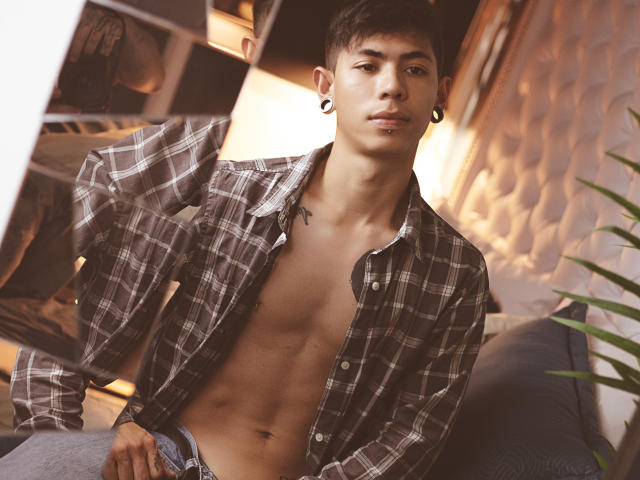 ArturCarter, shirtless, wearing unbuttoned plaid shirt, showing a toned, muscular chest, and a small tattoo near his collarbone, with black ear gauges and a confident, relaxed expression.