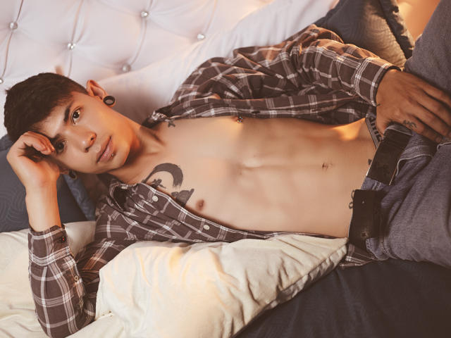 ArturCarter, shirtless, with a tattoo on his chest, lying on a bed, wearing an open plaid shirt, jeans, and ear gauges, looking at the camera, hand on his head.