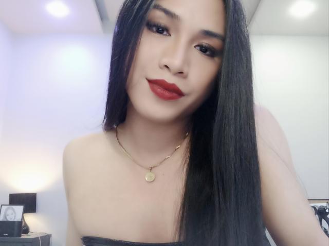 BarbaraFivo with long black hair, red lipstick, and a gold necklace, showing her bare shoulders and subtle cleavage, smiling softly at the camera.