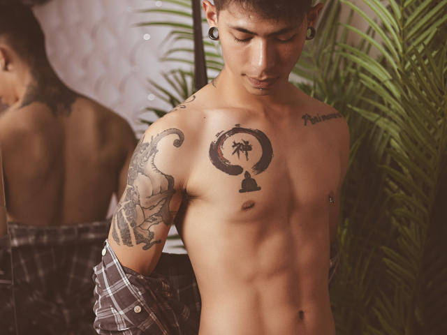 ArturCarter, shirtless, tan-skinned, tattooed, short brown hair, ear gauges, flannel off-shoulder, muscular, looking down, green plant, mirror reflection showing back.