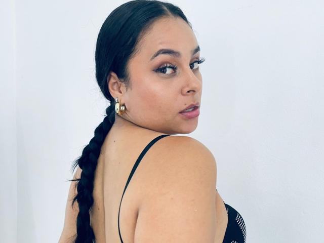 LunaSoler with long black braided hair, wearing a black spaghetti strap top, showing her bare back and small gold earrings, looking over her shoulder with a slight pout.