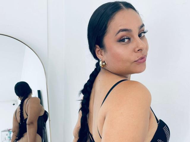 LunaSoler with a braided ponytail, black lingerie, and gold earrings, looking back with a pouty face, reflection in a mirror, showing her tits and ass.