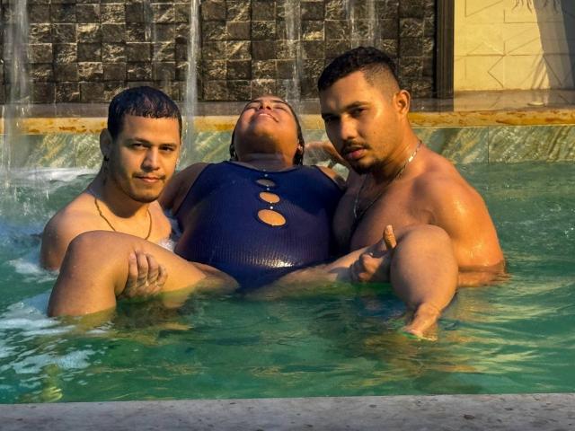 Three FantasticOrgyADs in a pool, one with a dark blue, cut-out one-piece swimsuit, surrounded by two shirtless FantasticOrgyADs, water splashing, sunlit, relaxed, muscular, attractive.