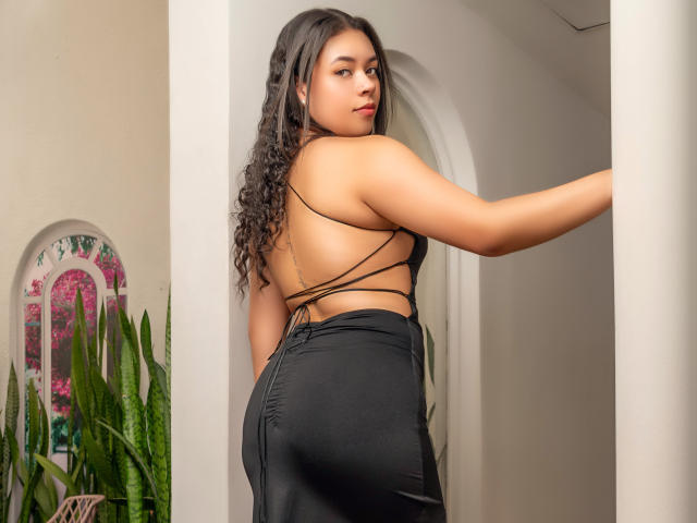 LexaStorm with curly black hair, wearing a black strappy dress, showing her back and ass, looking over her shoulder with red lipstick, standing near a white archway.
