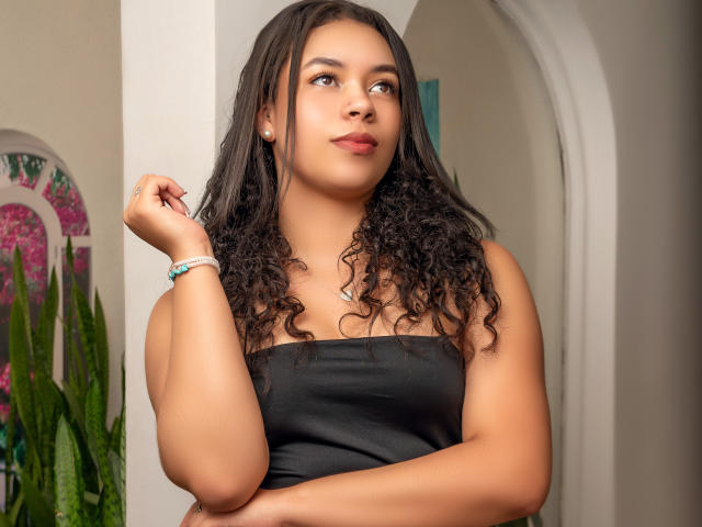 LexaStorm with curly black hair, wearing a black strap top, looking to the side with a slight smile, holding a lock of hair, tan skin, small earrings, and a bracelet.