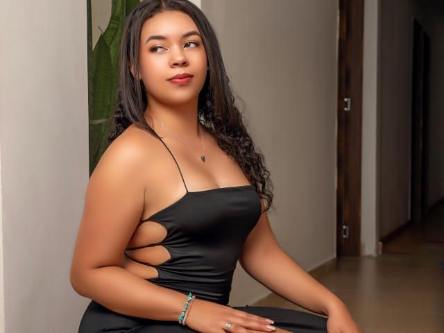 LexaStorm has tan skin, curly black hair, and is wearing a black cut-out dress, showing sideboob, with a silver necklace and bracelet, sitting and looking to the side.