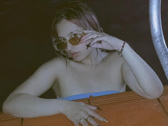 ElisaKim with wet brown hair, wearing small round sunglasses, a blue strapless top, and rings, leaning on an orange surface, hand near mouth, looking slightly bored.