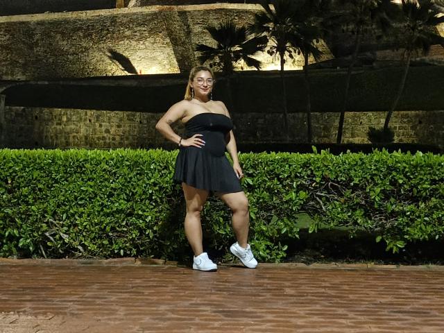 FantasykLadies with big ass, standing in black strapless dress, white sneakers, and hand on hip, smiling, in front of green hedge and brick path at night.