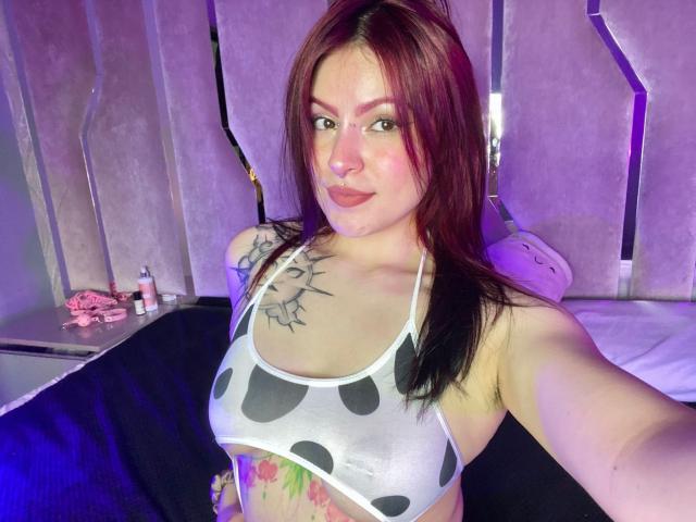 LilithHellsing with red hair, nose ring, and tattoos, wearing a cow print bikini top, showing her tits and colorful belly tattoo, taking a selfie.