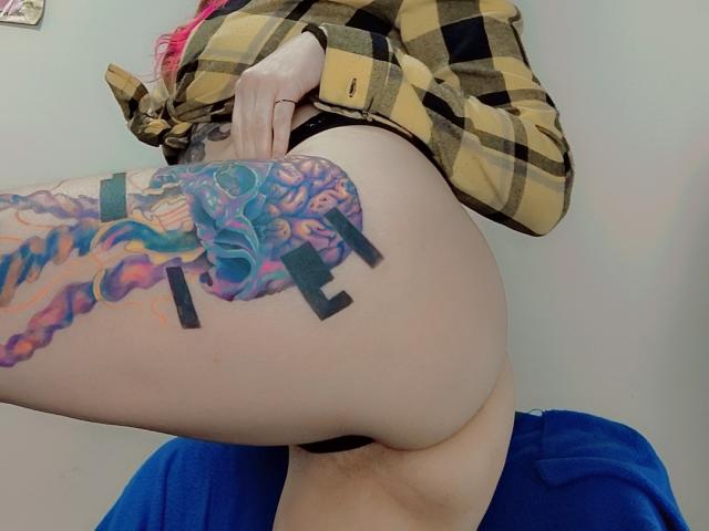 NatashaLau's bare ass, showing a colorful, intricate tattoo on her left thigh, wearing a yellow plaid shirt and blue pants, with pink hair visible.