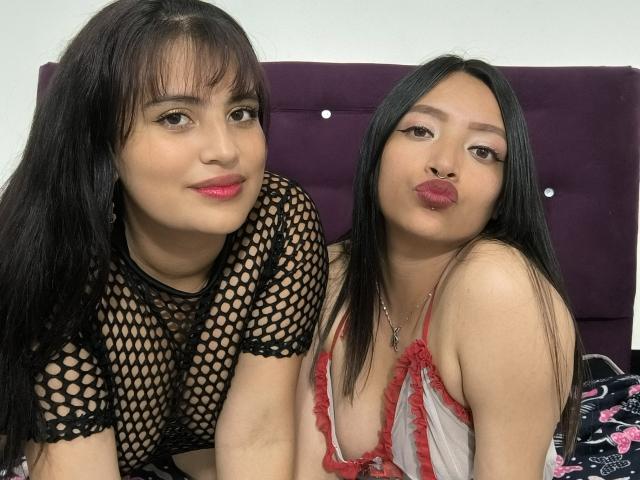 Two LesbiansMsi with long black hair, wearing fishnet and red lace tops, pouting with pink lipstick, sitting on a purple bed, showing their tits, smiling at the camera.
