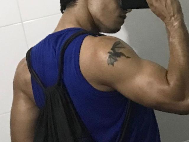 DaveBakers, muscular, dark-skinned, wearing a blue and black sleeveless shirt, taking a mirror selfie, showing a bird tattoo on his right bicep, holding a black phone.