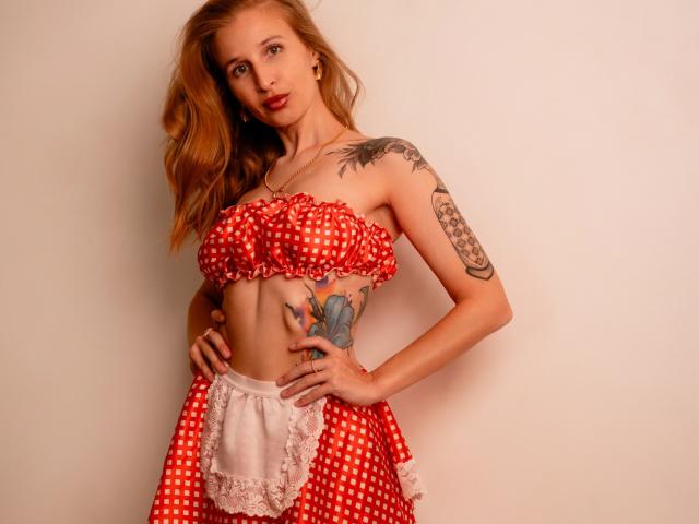 Nympheexe with red polka dot strapless top, showing her small tits, and matching skirt with white lace. She has tattoos on her arm and hip, standing with hands on hips.