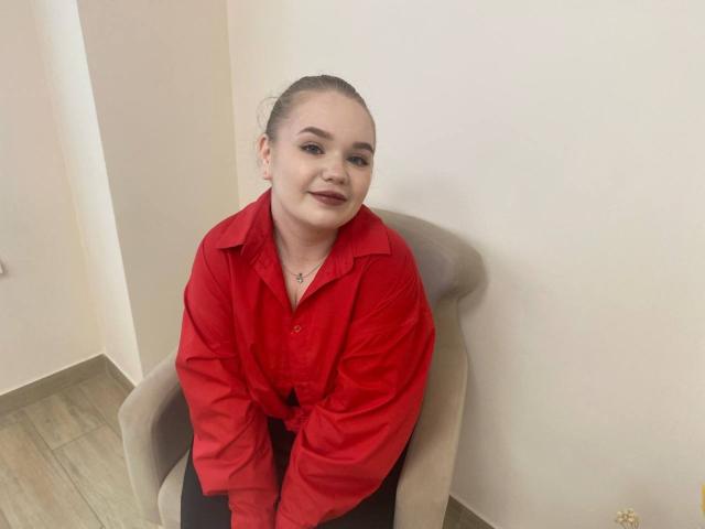EmmyWatsent, chubby with a red button-up shirt, sitting in a grey chair, has a cross necklace, dark red lipstick, and her hair is pulled back.