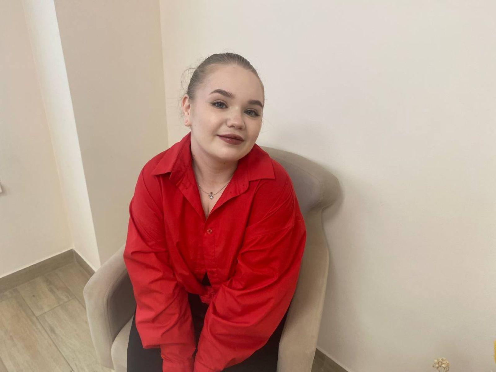 EmmyWatsent, chubby with a red button-up shirt, sitting in a grey chair, has a cross necklace, dark red lipstick, and her hair is pulled back.