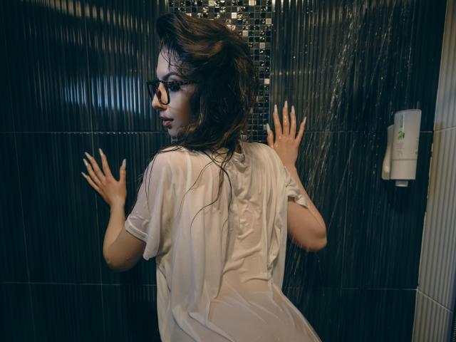 LucyGrace with wet, see-through shirt, hands against dark tiled wall, looking back over shoulder. She has long, wet brown hair and wears glasses. Her ass is slightly visible.