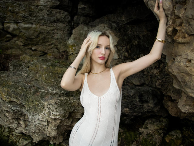 LexyGlam mit blonden Haaren, weißem Kleid und goldener Armband, steht vor einem felsigen Hintergrund, der rechte Arm ist erhoben, die linke Hand im Haar, sie zeigt ihre Brüste und ihren Hintern, rosa Lippen, selbstbewusst..