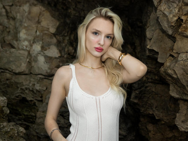 LexyGlam with blonde hair, pink lipstick, white knit tank top, gold bracelet, and necklace, standing against rocky background, hand on cheek, looking at camera, tits visible.