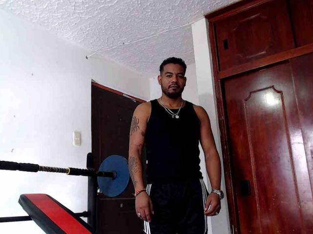 BladeFire, muscular, dark-haired, bearded, tattooed arms, wearing black tank top and black shorts, standing in front of wooden door, looking serious, with weight bench in background.