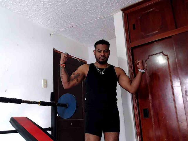 BladeFire, muscular, black tank top, black shorts, tattooed arms, flexing, standing in front of wooden door, ring on finger, wristband, black weight bench with blue weight plate.