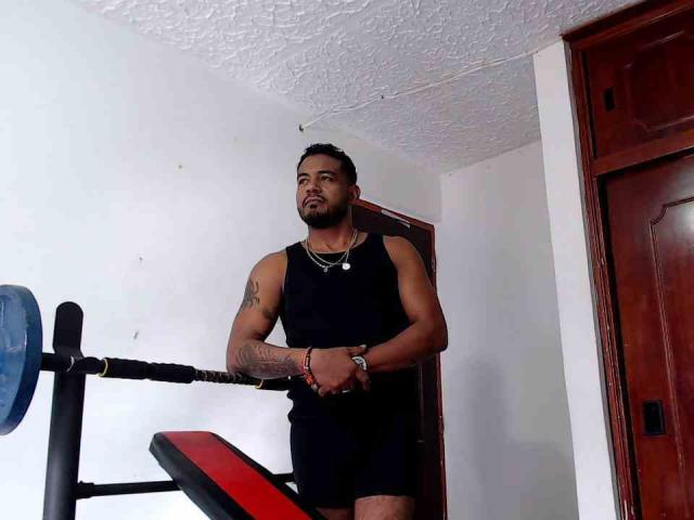 BladeFire, muscular Latino dude with tattoos, black tank top, and shorts, standing near a red and black weight bench, looking serious, in a white-walled room with wooden doors.