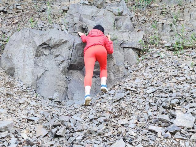 Stellina1-hot, butt in red pants, lying face-down on rocky ground, wearing red jacket, black cap, white socks, blue shoes, arms stretched forward, surrounded by rocks and dirt.