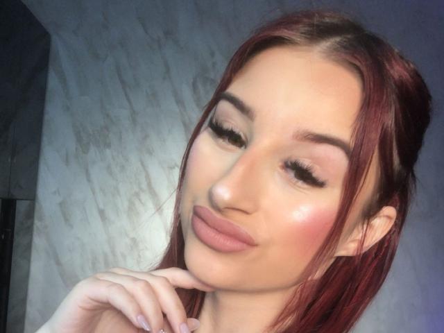 FoxxyAmber with red hair, closed eyes, and pouty lips, has long eyelashes, subtle pink blush, and is touching her chin with a hand, showing manicured nails.